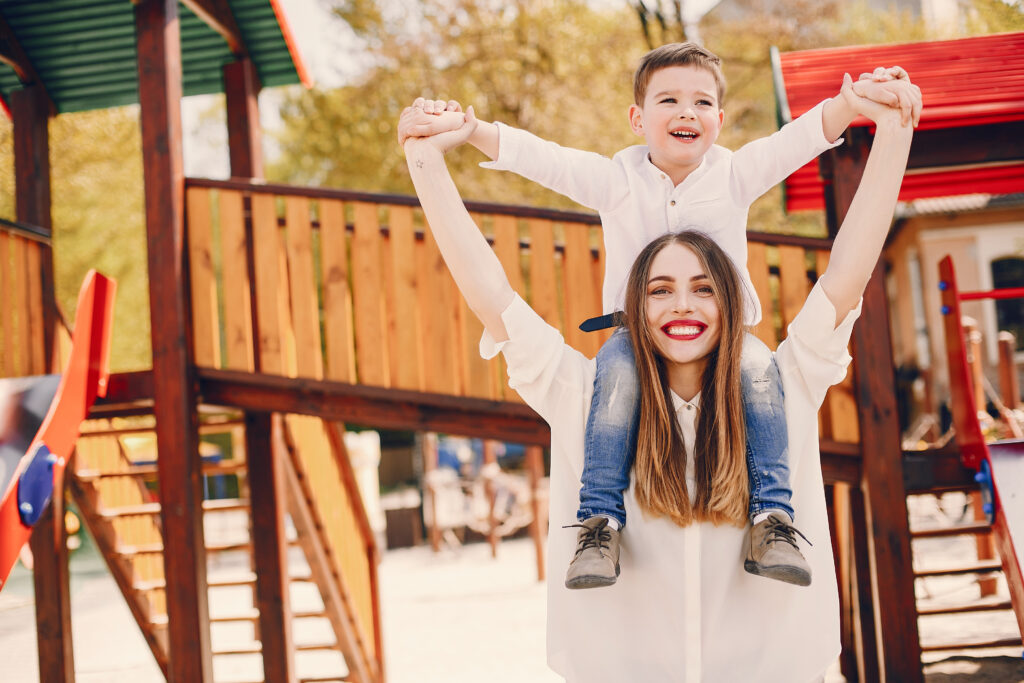 giwa-mamma-con-bimbo-sulle-spalle-e-sullo-sfondo-una-casetta-in-legno-di-un-parco-giochi